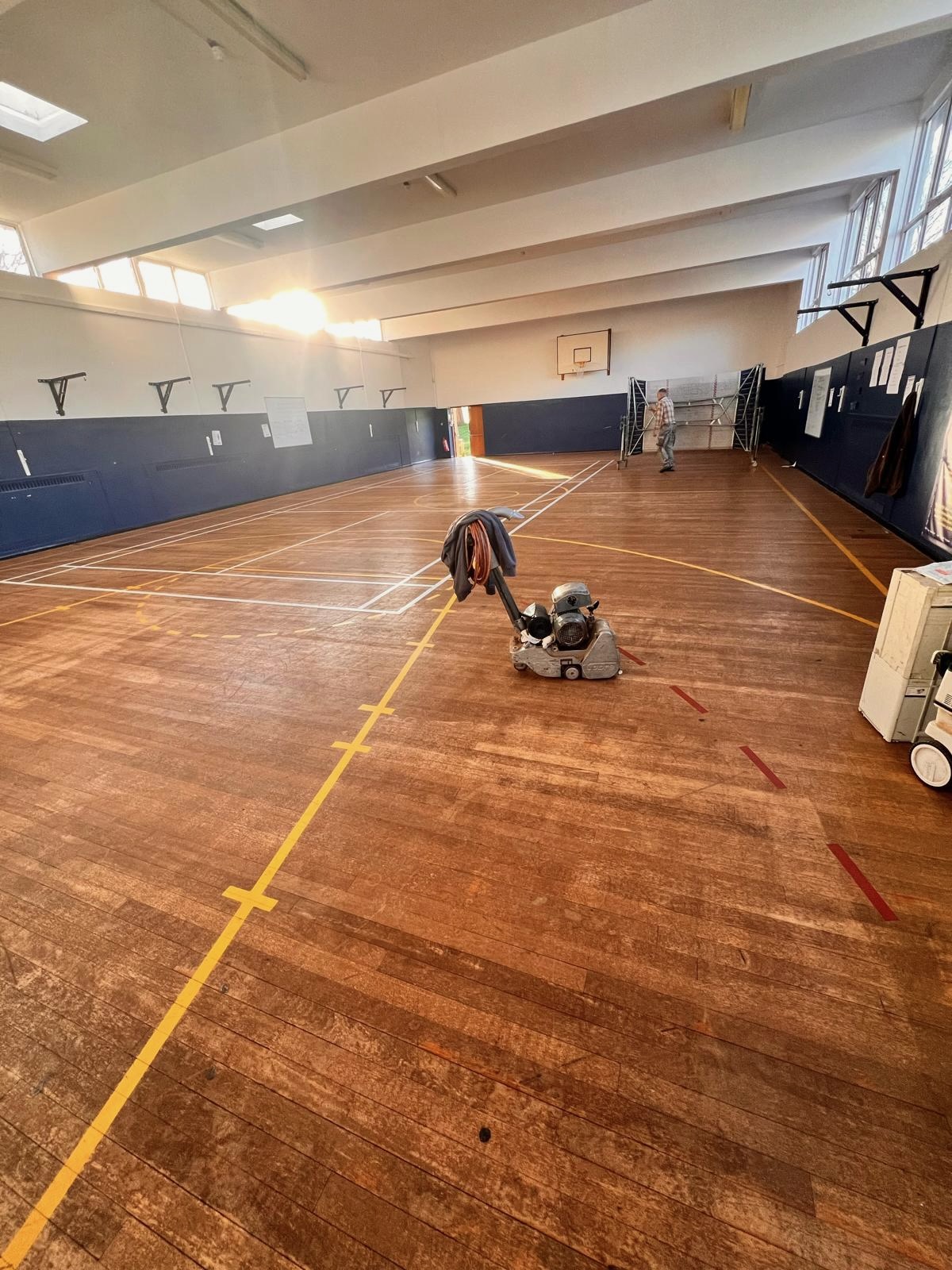 Belt sander removing worn yellow and red court markings from a school sports hall floor